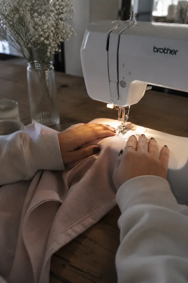 Hands guiding blush-pink fabric through a sewing machine on a wooden table, with a jar of dried flowers beside it.